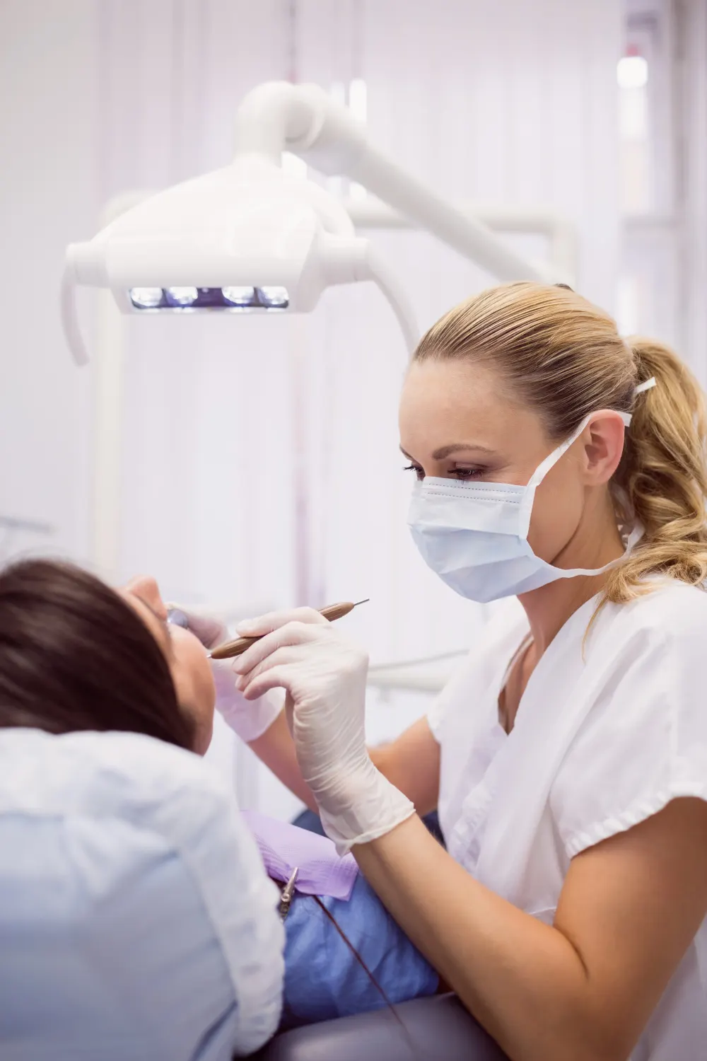 A clinician in a face mask and gloves examines a patient's teeth under a dental treatment light